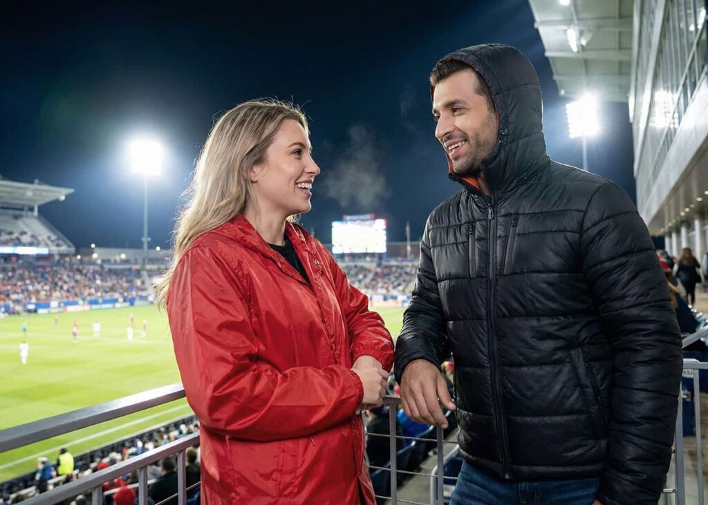 woman in red coach jacket and man in hooded puffer jacket watching night sports game in stands