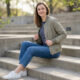 woman sitting on outdoor steps wearing a lightweight bomber jacket striped top jeans and white sneakers