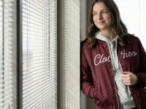 woman in a maroon Clothoo bomber jacket layered over a gray hoodie standing by an office window