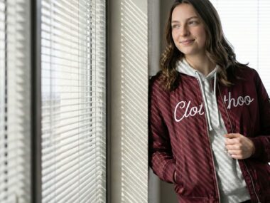 woman in a maroon Clothoo bomber jacket layered over a gray hoodie standing by an office window